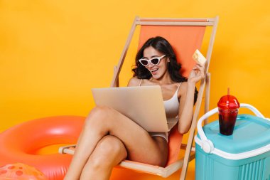 Sarı duvarda izole edilmiş şezlongda dinlenirken elinde kredi kartı olan mutlu bir kadın görüntüsü.