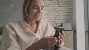 Akıllı telefon kullanan ve evdeki kanepede otururken pencereye bakan mutlu, çekici sarışın kadın.