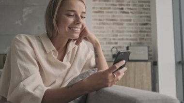 Evdeki kanepede otururken akıllı telefon kullanan neşeli, çekici sarışın kadın.