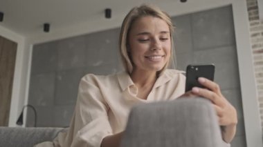 Evdeki kanepede otururken akıllı telefon kullanan çekici sarışın kadın.