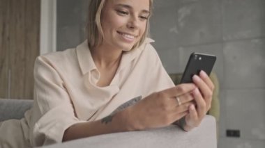 Evdeki kanepede otururken akıllı telefon kullanan neşeli, çekici sarışın kadın.
