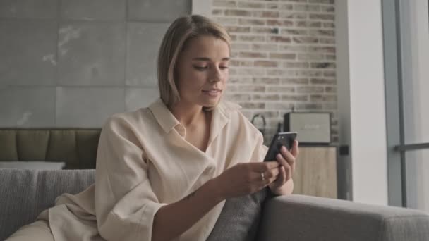 Mujer Rubia Atractiva Sonriente Usando Teléfono Inteligente Mientras Sienta Sofá — Vídeos de Stock