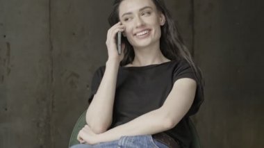 Neşeli, güzel esmer kadın akıllı telefonla konuşuyor ve gri duvarın üzerinde otururken kafasını çeviriyor.