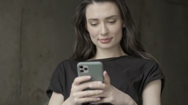 Gri duvardaki sandalyede otururken akıllı telefondan mesaj yazan konsantre olmuş esmer kadın.