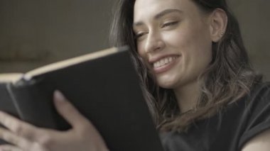 Neşeli, güzel esmer kadının kitap okurken gri duvarın üzerinde oturduğu manzarayı kapat.