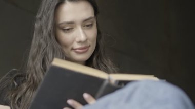 Sakin, güzel esmer kadın, gri duvarın üzerinde otururken kitap okuyor.