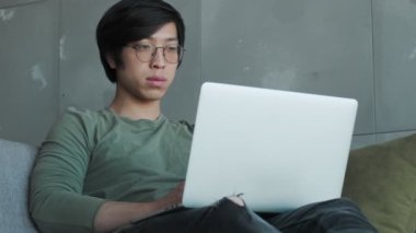 Gözlüklü genç Asyalı bir adam dairede koltukta otururken dizüstü bilgisayarını kullanıyor.