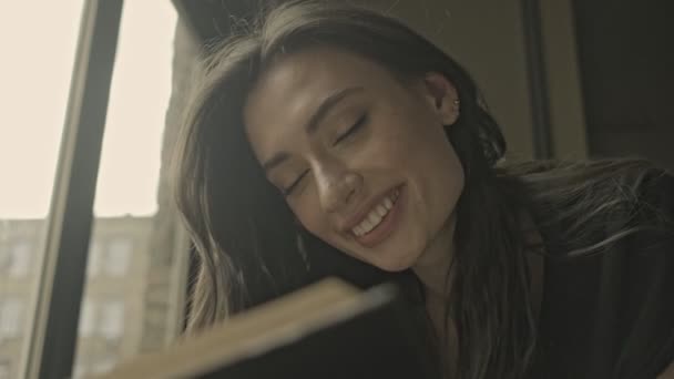 Усміхнена Красива Брюнетка Читає Книгу Потім Дивиться Вікно Лежачи Підлозі — стокове відео