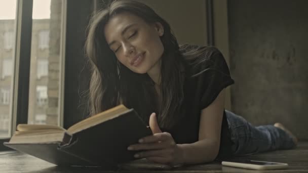 ハッピー可愛いですブルネット女性読書本を読みながら床の上に寝そべって屋内 — ストック動画