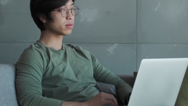 一个戴眼镜的有思想的亚洲年轻人正坐在公寓的沙发上 一边用笔记本电脑 — 图库视频影像