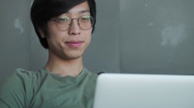 Mutlu Asyalı gözlüklü genç bir adam evinde koltukta otururken dizüstü bilgisayarını kullanıyor.