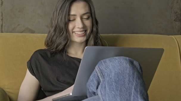 Jolie jolie femme brune utilisant un ordinateur portable puis regardant loin tout en étant assis sur le canapé à l'intérieur