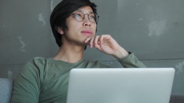 Gözlüklü yakışıklı genç Asyalı bir adam dizüstü bilgisayarıyla çalışırken bir şeyler hayal ediyor.