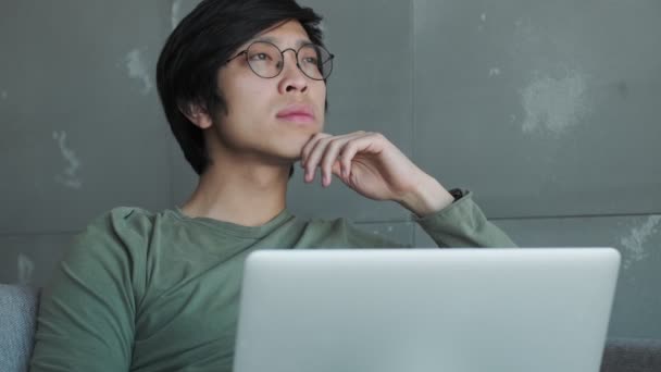 Симпатичный Молодой Азиат Очках Думает Пока Работает Своим Ноутбуком Сидящим — стоковое видео