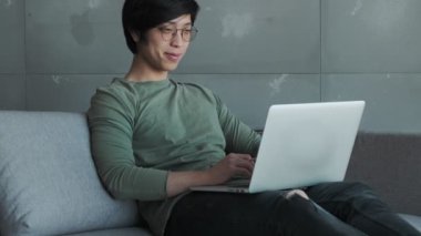 Gözlüklü, gülümseyen Asyalı bir adam dizüstü bilgisayarıyla çalışıyor. Dairedeki koltukta oturuyor.