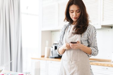 Modern mutfakta turta pişirirken cep telefonuyla önlük giyen esmer kadın resmi