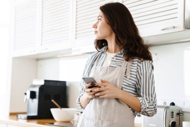 Modern mutfakta turta pişirirken cep telefonuyla önlük giyen esmer kadın resmi