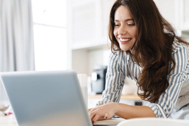 Modern mutfakta turta pişirirken dizüstü bilgisayar kullanan esmer bir kadın resmi