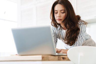 Modern mutfakta turta pişirirken dizüstü bilgisayar kullanan esmer bir kadın.