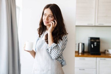 Modern mutfakta kahve içerken cep telefonuyla konuşan önlük giymiş mutlu bir kadın görüntüsü.
