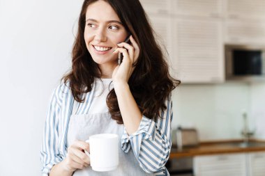 Modern mutfakta kahve içerken cep telefonuyla konuşan önlük giymiş mutlu bir kadın görüntüsü.