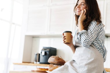 Modern mutfakta kahve içerken cep telefonuyla konuşan önlük giymiş mutlu bir fırıncı kadın resmi.
