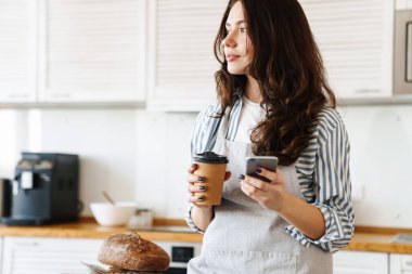 Modern mutfakta kahve içerken önlük takan güzel bir fırıncı kadın resmi.