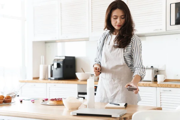 Image of focused brunette woman wearing apron using cellphone while cooking pie in modern kitchen