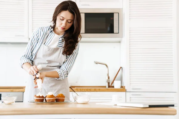 イメージのCaucasianブルネット女性身に着けていますエプロン調理マフィンとともにクリームで現代的なキッチン — ストック写真