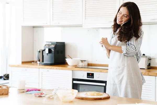 Image of laughing beautiful woman wearing apron talking on cellphone while cooking pie in modern kitchen