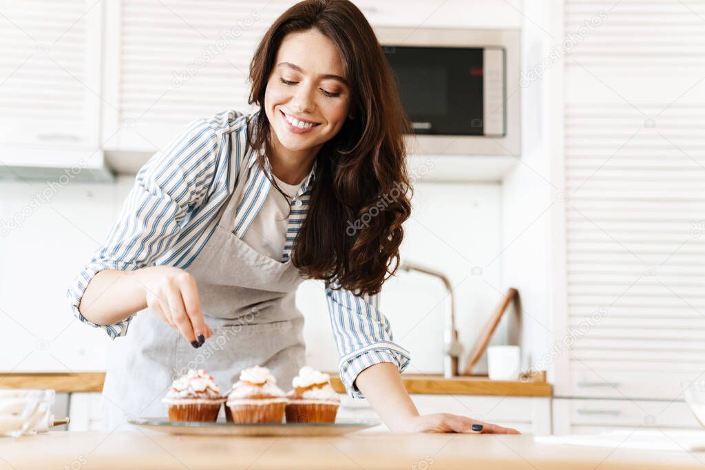Image of caucasian happy woman wearing apron smiling and cooking muffins in modern kitchen