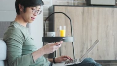 Gözlüklü yakışıklı bir Asyalı 'nın yan görüntüsü, evinde oturma odasında dizüstü bilgisayarıyla çalışırken akıllı telefonunu kullanıyor.