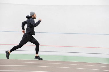 Dışarıdaki stadyumdaki yarış pistinde koşan genç ve formda bir sporcunun yan görüntüsü.