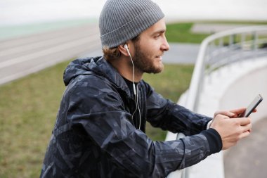 Motivasyonlu genç sporcu kulaklıkla müzik dinlerken stadyumda duruyor, cep telefonu kullanıyor, demiryoluna yaslanıyor.