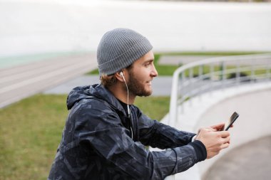 Motivasyonlu genç sporcu kulaklıkla müzik dinlerken stadyumda duruyor, cep telefonu kullanıyor, demiryoluna yaslanıyor.