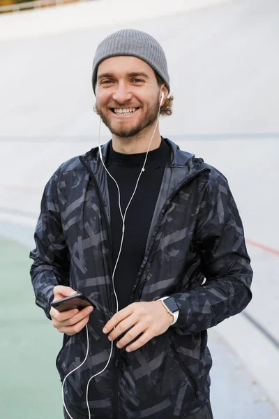 Motywowany Uśmiechnięty Młody Wysportowany Sportowiec Słuchający Muzyki Słuchawek Stojąc Stadionie — Zdjęcie stockowe