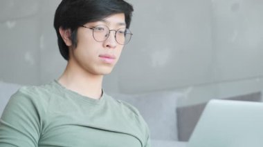 Gözlüklü yakışıklı Asyalı bir adamın yakın plan görüntüsü dizüstü bilgisayarını evdeki oturma odasında oturuyor.