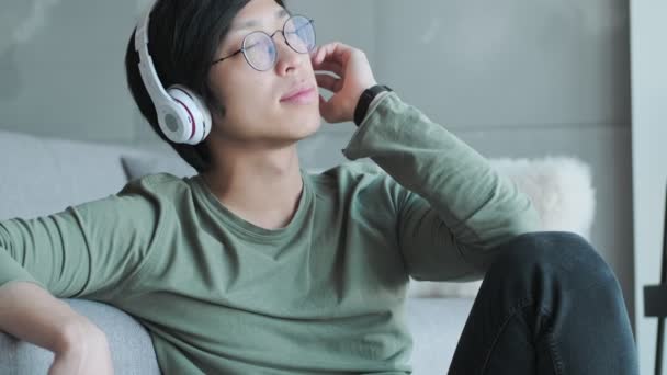 自宅のリビングルームに座って音楽を楽しんだヘッドフォンを持つリラックスした若いアジアの男 — ストック動画