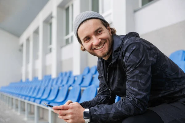 Uśmiechnięty Młody Sportowiec Siedzący Trybunach Stadionie Korzystający Telefonu Komórkowego — Zdjęcie stockowe