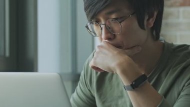 Gözlüklü, odaklanmış, genç Asyalı bir bireyin yakın plan görüntüsü evde masada otururken dizüstü bilgisayarını kullanıyor.