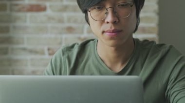 Gözlük takan konsantre olmuş Asyalı genç bir bireyin yakın plan görüntüsü evde masada otururken dizüstü bilgisayarını kullanıyor.