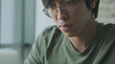 Gözlük takan konsantre olmuş Asyalı genç bir bireyin yakın plan görüntüsü evde masada otururken dizüstü bilgisayarını kullanıyor.