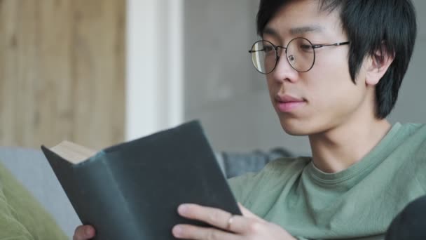 Красивий Молодий Азіатський Чоловік Окулярах Читає Книгу Сидячи Вітальні Вдома — стокове відео