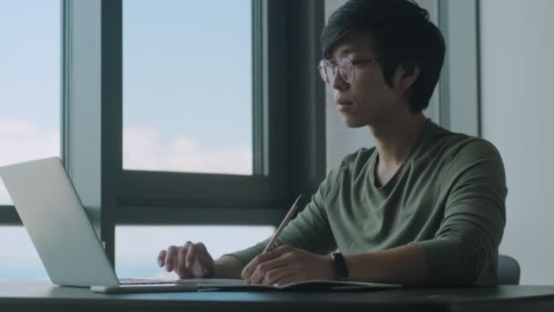 一位戴着眼镜的年轻的亚洲男子在家中与笔记本电脑一起工作时正在做笔记 — 图库视频影像