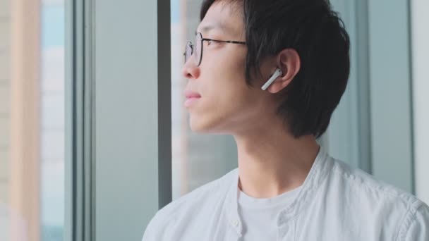 Ένας Γοητευτικός Νεαρός Ασιάτης Ακουστικά Κοιτάζει Έξω Από Παράθυρο Στο — Αρχείο Βίντεο