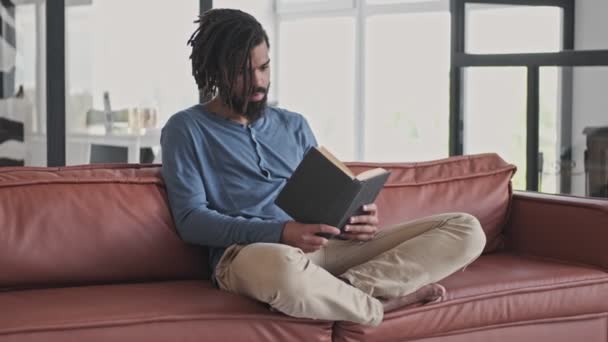 Glad Afrikansk Man Vardagskläder Läser Bok Sittandes Soffan Vardagsrummet Hemma — Stockvideo