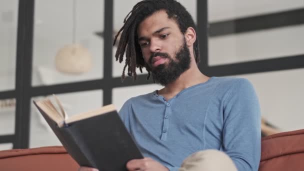一位心情愉快的积极的非洲裔美国人穿着休闲装 正坐在客厅的沙发上看书 — 图库视频影像