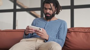 Ciddi bir Afrikalı genç Amerikalı adam evdeki oturma odasında akıllı telefondan oyun oynuyor.