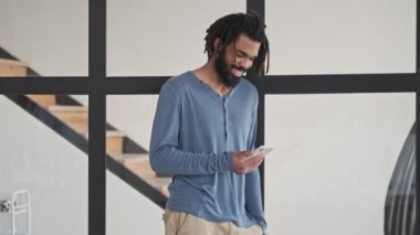 Gülümseyen Afro-Amerikan bir adam akıllı telefonunu evdeki oturma odasında kullanıyor.