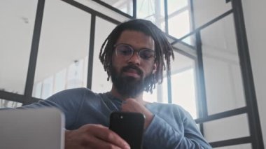 Odaklanmış Afro-Amerikan bir adam evinde oturma odasında laptopuyla çalışırken akıllı telefon kullanıyor.